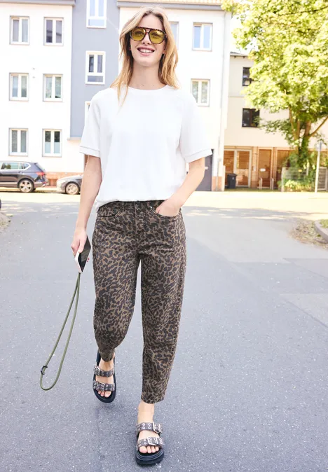 Femme en t-shirt blanc, pantalon à motifs et sandales se promène par une rue résidentielle ensoleillée.