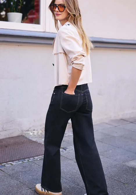 Femme en veste claire et lunettes de soleil porte un jean noir ample, debout sur le trottoir, regarde par-dessus son épaule.