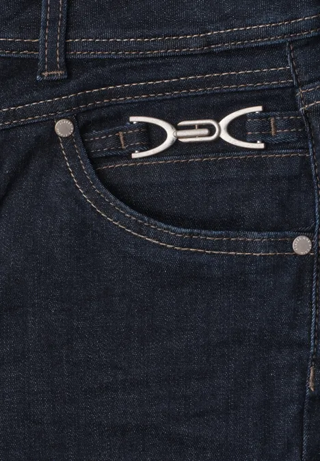 Gros plan d'un jean bleu foncé, poche avant ornée d'un logo métallique argenté en forme de double arc.