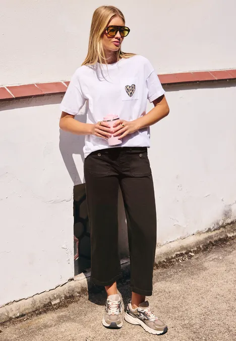 Blonde vrouw met zonnebril, wit T-shirt en zwarte broek staat ontspannen voor een witte muur.