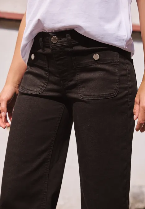 Close-up van een persoon in zwarte high-waist jeans en wit shirt, handen ontspannen langs het lichaam