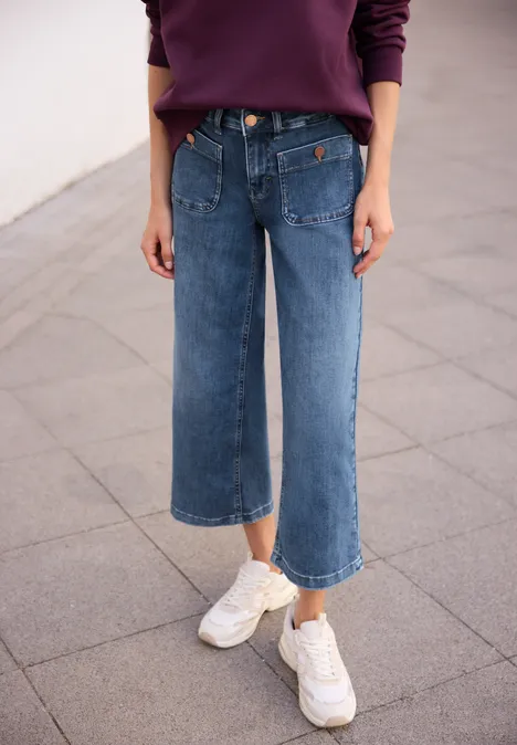 Mid Waist Wide Leg Jeans im Loose Fit authentic mid blue wash Person in weitem, knöchellangen Jeans und weißen Sneakern auf Betonboden.
