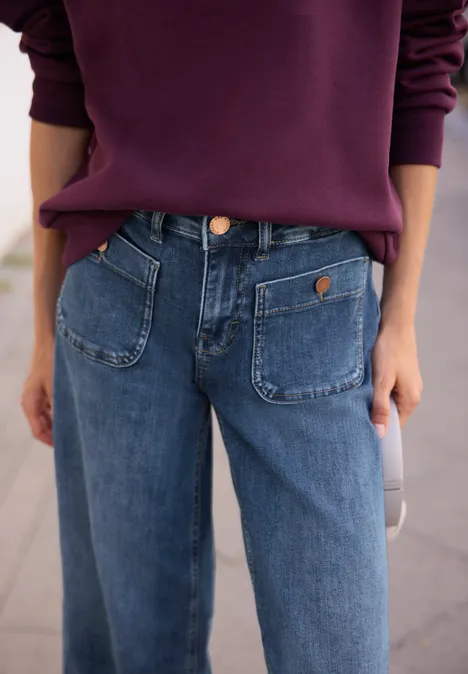 Mid Waist Wide Leg Jeans im Loose Fit authentic mid blue wash Nahaufnahme einer Person in blauen Jeans mit auffälligen Taschen und lila Pullover.