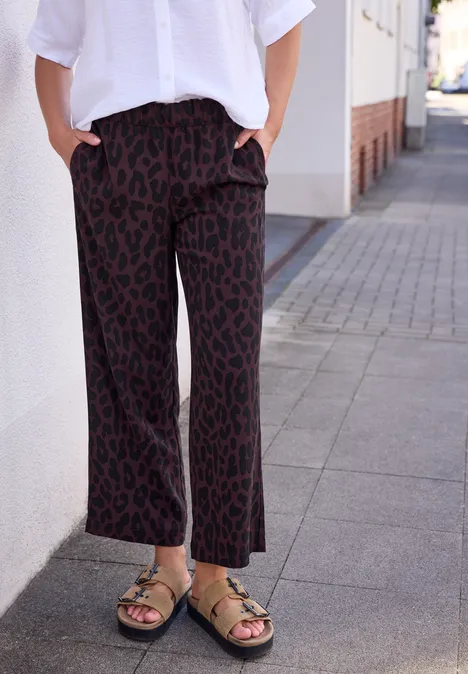 Personne portant un pantalon ample à motif, un haut blanc et des sandales vertes, adossée à un mur