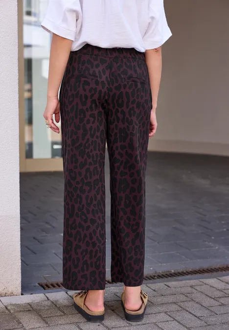 Une personne vue de dos porte un pantalon large à motif léopard rouge et des sandales dans un cadre urbain.