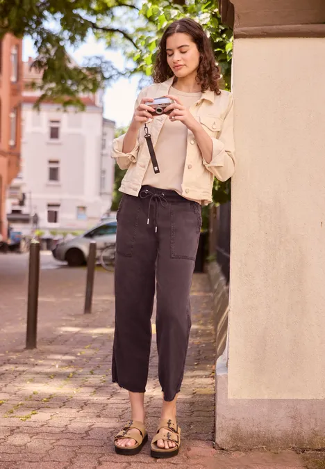 Een vrouw in lichte blouse en sandalen leunt buiten tegen een gebouw en kijkt op haar smartphone.