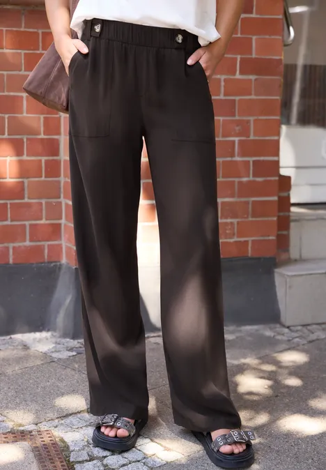 Pantalon noir ample droit et sandales noires portés devant un mur en briques.