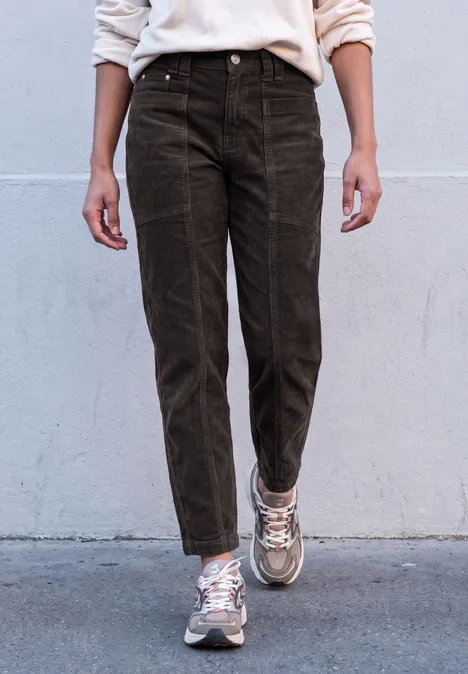 Eine Person trägt braune Cordhose und weiße Sneakers vor einer grauen Wand.