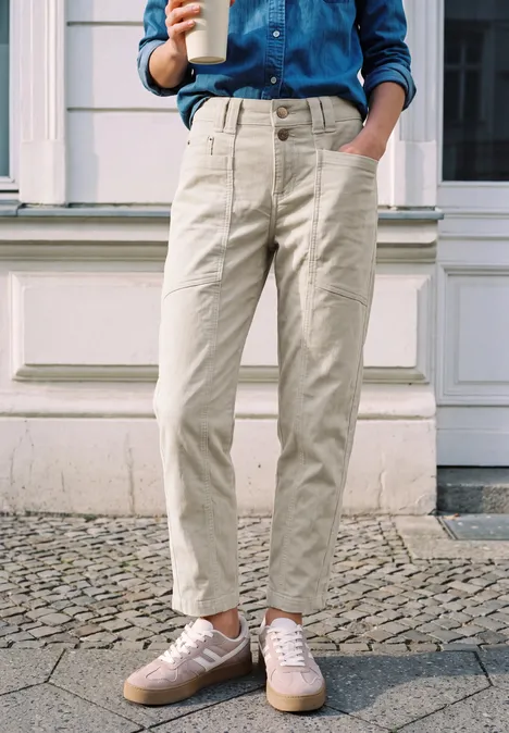 Personne debout sur un trottoir pavé, porte un pantalon beige, une chemise en jean, des baskets et tient un café.