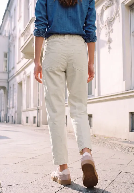 Une personne de dos, en pantalon clair et veste en jean, marche dans une rue ensoleillée.