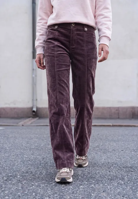 Personne de la taille aux pieds portant un pantalon velours côtelé violet et des baskets, marchant sur l'asphalte devant un mur.