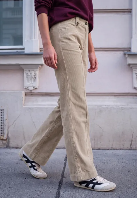 Personne en pantalon velours beige clair et baskets blanches marche devant une façade.