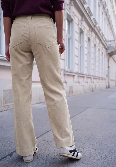 Personne en pantalon en velours beige et baskets pose sur un trottoir devant une façade classique.
