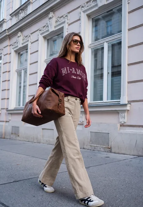Une femme élégante en pantalon beige, sweat bordeaux et lunettes de soleil marche près d’une façade