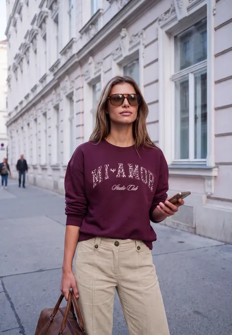 Femme en lunettes de soleil, sweat bordeaux 'Mi AMOR', pantalon beige, sac et téléphone, marchant dans une rue urbaine.