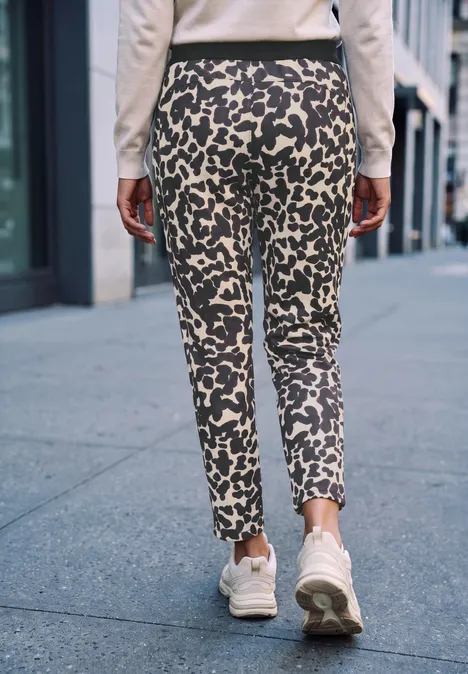 Eine Person geht auf einem Gehweg, trägt enge, gemusterte Hose mit Leopardenflecken und weiße Sneaker.