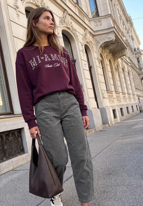 Eine Frau mit burgunderrotem Pullover, grauen Hosen und Ledertasche spaziert vor klassischer Stadtkulisse.