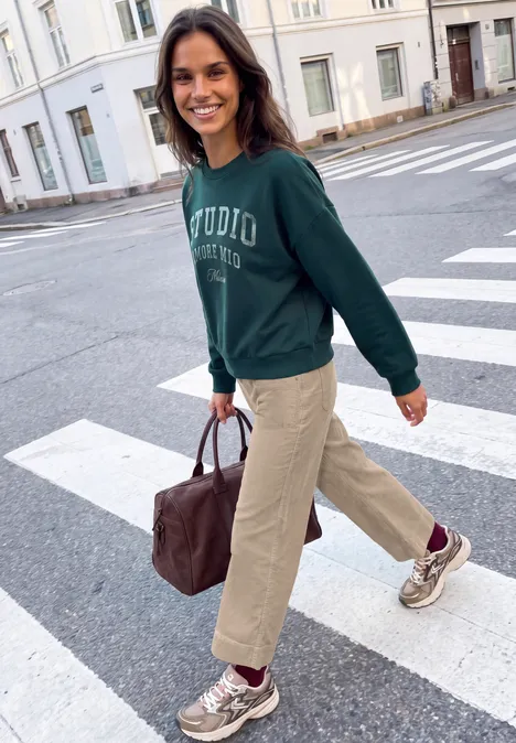 Mid Waist Wide Leg Broek – Casual Fit biscuit sand Frau in grünem Pullover und Tasche überquert lächelnd Zebrastreifen in der Stadt.