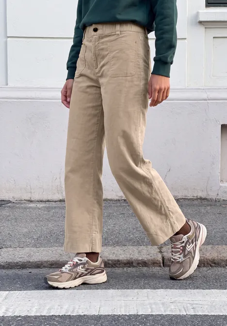 Mid Waist Wide Leg Broek – Casual Fit biscuit sand Person geht über Zebrastreifen, trägt beige weite Hose, gemusterte Sneaker und grünen Pullover.