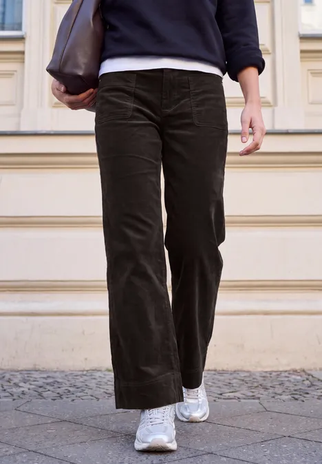 Person in dunkelgrüner Cordhose, weißen Sneakern und brauner Ledertasche vor einer Hausfassade.