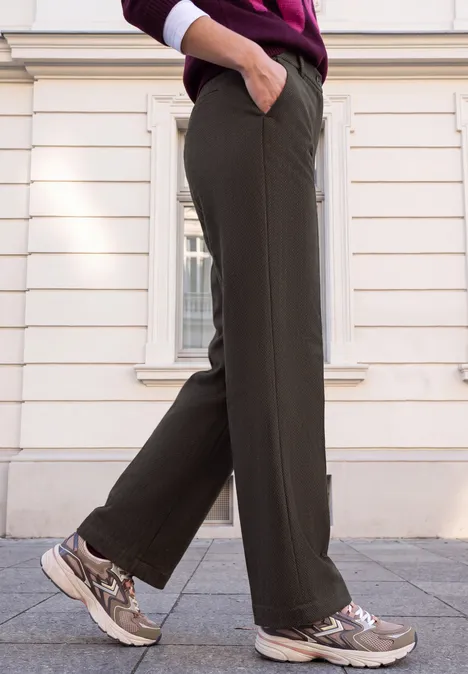 Personne de profil portant un pantalon olive et des baskets beiges, marchant sur des pavés devant une façade blanche