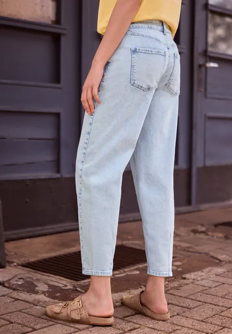 Rückenansicht einer Person in hellblauen Jeans und flachen Sandalen auf gepflastertem Gehweg.