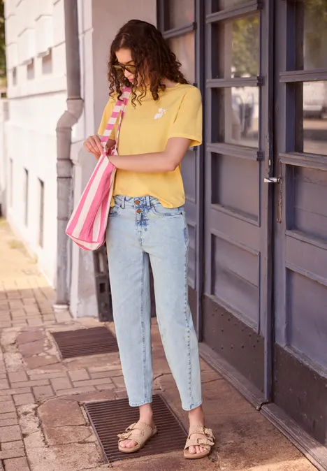 Frau mit gelbem T-Shirt, blauen Jeans und Sandalen steht mit rosafarbener Tasche vor einer Haustür.