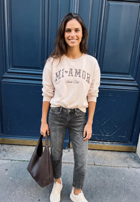Junge Frau in Jeans und cremefarbenem Sweatshirt mit Tasche posiert vor einer blauen Tür.