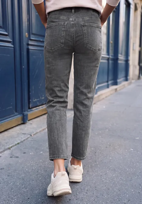 Person von hinten, trägt graue Jeans und weiße Sneaker, läuft auf Bürgersteig vor blauem Gebäude.