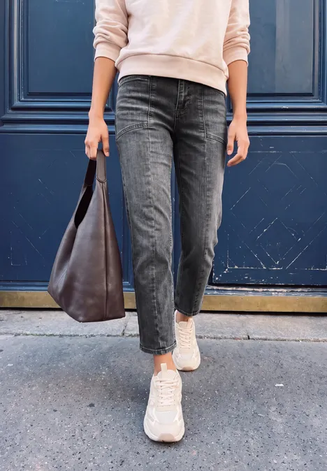 Person in schwarzen Jeans und weißen Sneakern trägt eine braune Ledertasche vor einer blauen Tür.
