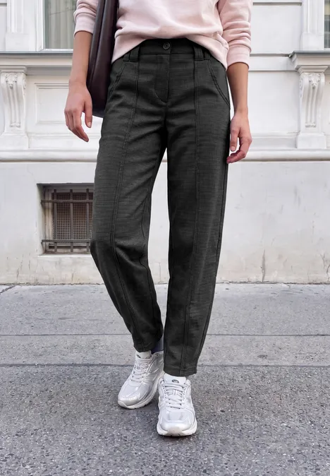 Person steht vor einem Gebäude und trägt schwarze Stoffhose mit weißen Sneakers auf Asphaltpflaster.