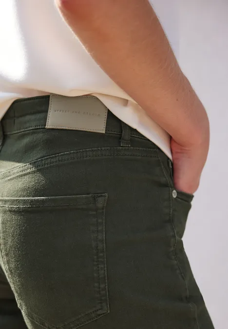 Rückansicht einer Person in olivgrünen Jeans und weißem Oberteil mit einer Hand in der Gesäßtasche.