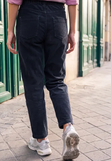 Person in schwarzen Jeans und weißen Sneakers von hinten auf einem Gehweg vor grünen Türen.