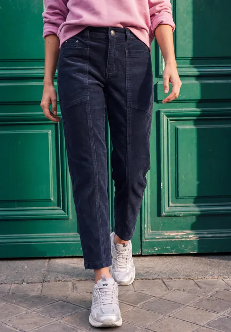 Person im rosa Pullover, schwarzer Jeans und weißen Sneakern steht vor einer grünen Tür.