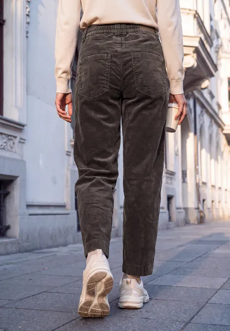 Mid Waist Barrel Leg Hose im Loose Fit charred brown Eine Person in Cordhosen und weißen Sneakern geht auf einem Gehweg zwischen Altbauten.