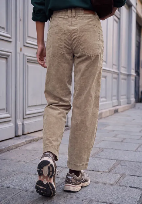 Mid Waist Barrel Leg Hose im Loose Fit henna brown Rückansicht einer Person in beigen Cordhose und Turnschuhen auf gepflastertem Gehweg vor einer Hausfassade.