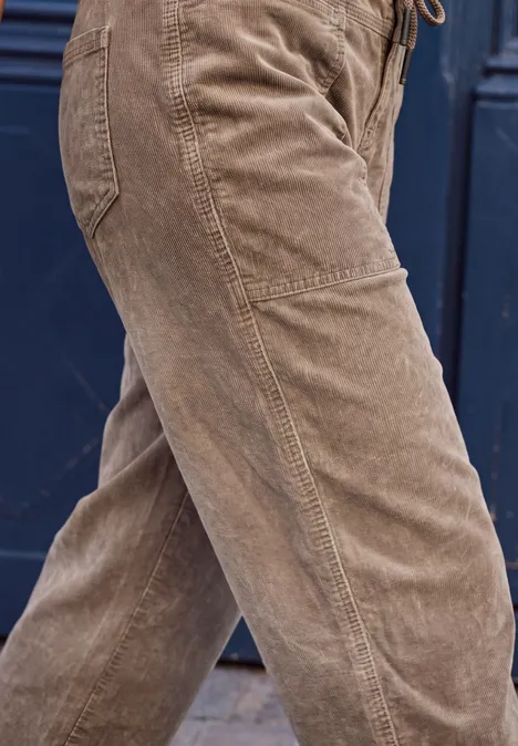 Mid Waist Barrel Leg Hose im Loose Fit henna brown Braune Cordhose an Personenbeinen, seitliche Nahaufnahme vor dunklem Hintergrund.