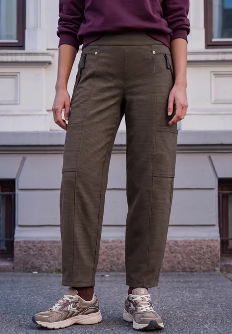 Person steht in olivgrüner Hose und Sneakers vor einem Gebäude.