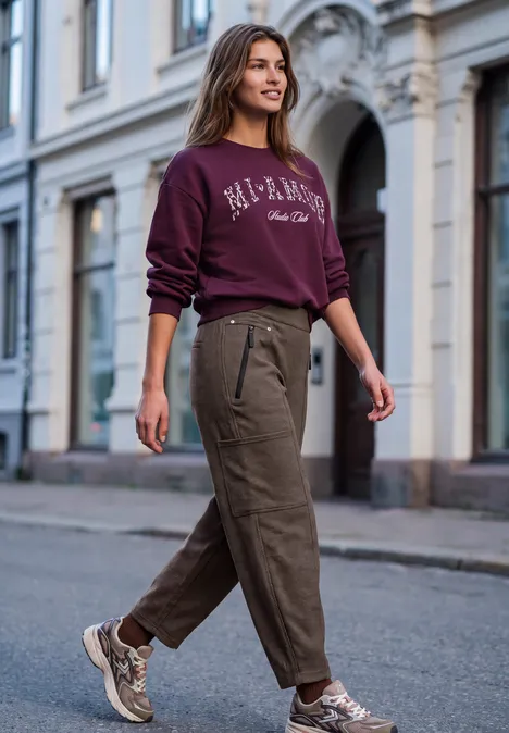Eine junge Frau läuft in City-Straßen, trägt bordeauxfarbenen Pullover, khakifarbene Hose und Sneakers.