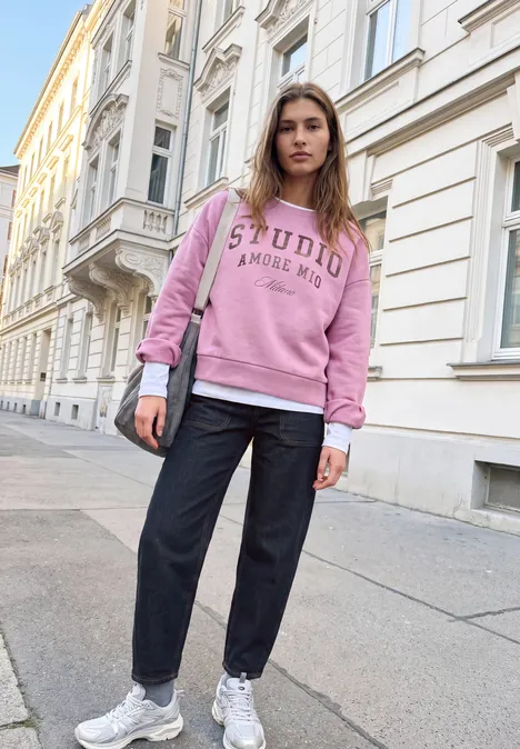 Relaxed Barrel Leg Leg Jeans im Loose Fit rinse washed Junge Frau in rosa Sweatshirt, schwarzer Hose und weißen Sneakern steht auf Gehsteig vor Altbau.
