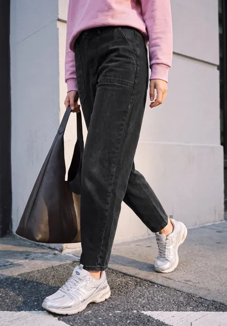 Relaxed Barrel Leg Leg Jeans im Loose Fit soft black washed Person in schwarzer Cordhose, rosa Pullover und weißen Sneakern trägt schwarze Tasche und geht über Gehweg.