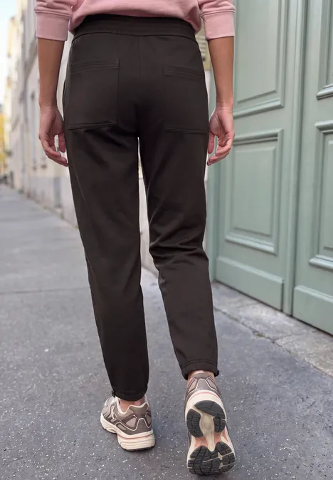 Mid Waist Barrel Leg Hose im Loose Fit charred brown Rückenansicht einer Person in schwarzen Hosen und Sneakern, die auf einem Bürgersteig läuft.