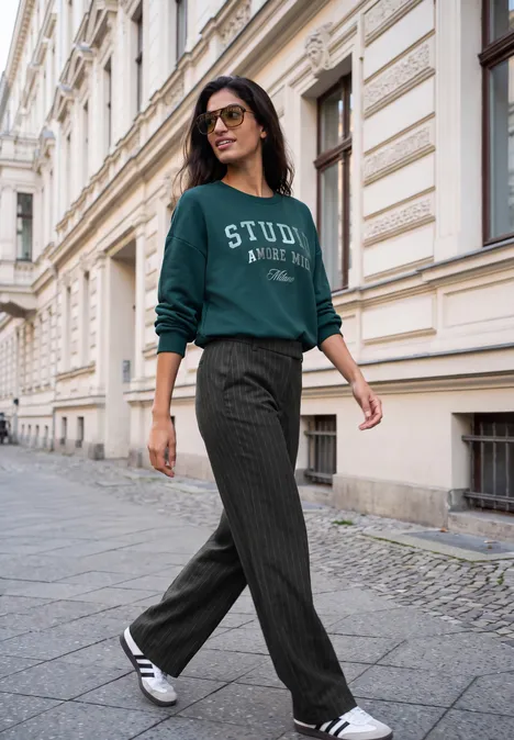 High Waist Straight Leg Hose im Casual Fit Black Eine Frau mit Brille und grünem Sweatshirt läuft in lässiger Hose und Sneakern an historischer Straße entlang.