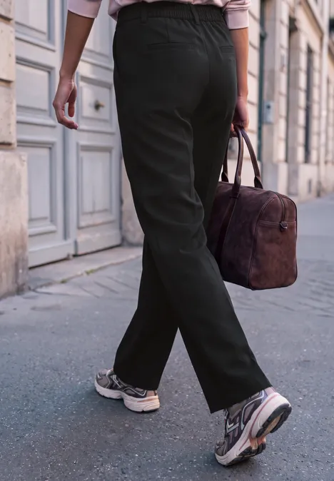 High Waist Straight Leg Hose im Casual Fit Black Eine Person mit schwarzen Hosen und Sneakern läuft auf dem Gehweg und hält eine braune Tasche.
