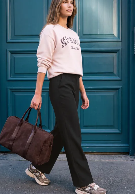 High Waist Straight Leg Hose im Casual Fit Black Frau in lässigem Pullover und schwarzen Hosen trägt eine braune Reisetasche vor einer türkisfarbenen Haustür.