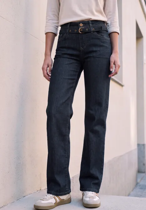 Personne porte un jean foncé, un haut blanc et des baskets blanches contre un mur.