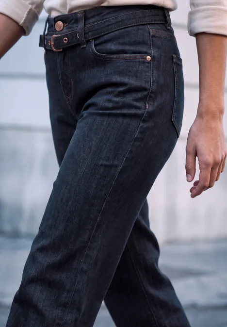 Vue partielle d’une personne en jean bleu foncé avec ceinture, debout devant un arrière-plan flou.