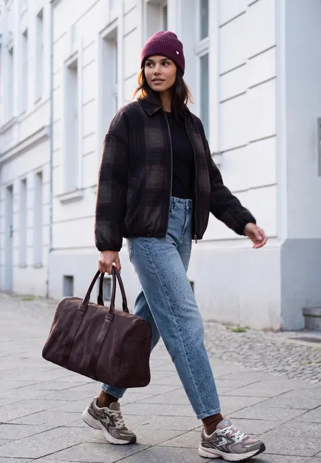Vrouw in jeans, jas en muts loopt op een stadsstoep en draagt een reistas.