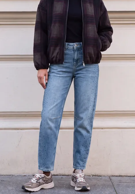 Persoon met paarse geruite bomber, zwarte trui, blauwe jeans en grijze sneakers voor een gevel.