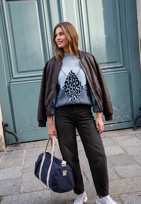 Jeans Relaxed Barrel Leg o kroju Loose Fit black washed Młoda kobieta przed niebieskimi drzwiami, skórzana kurtka na ramionach, wzorzysty sweter, czarne spodnie i torba.